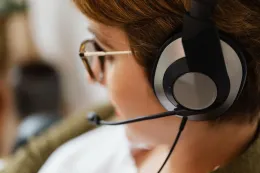 A detailed, candid close-up photograph of a professional woman in profile, wearing a high-quality black over-ear headset with a prominent silver ear cup plate and an integrated adjustable boom microphone extending toward her mouth.