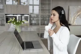 Medium shot of a woman working from home on a laptop, participating in a video conference, wearing a headset in a bright, professional home office.