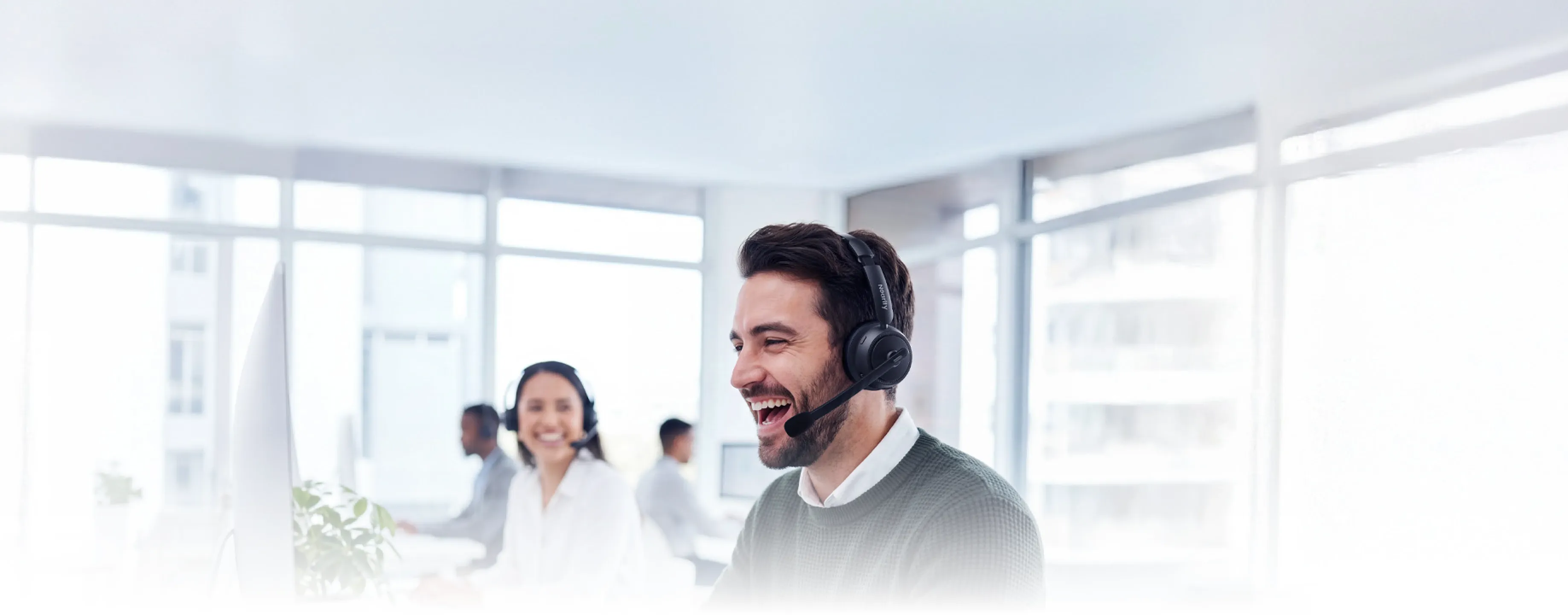 A smiling professional in a busy workspace wearing a noise cancelling headphone with mic for office, featuring ProperClean AI environmental noise cancellation.