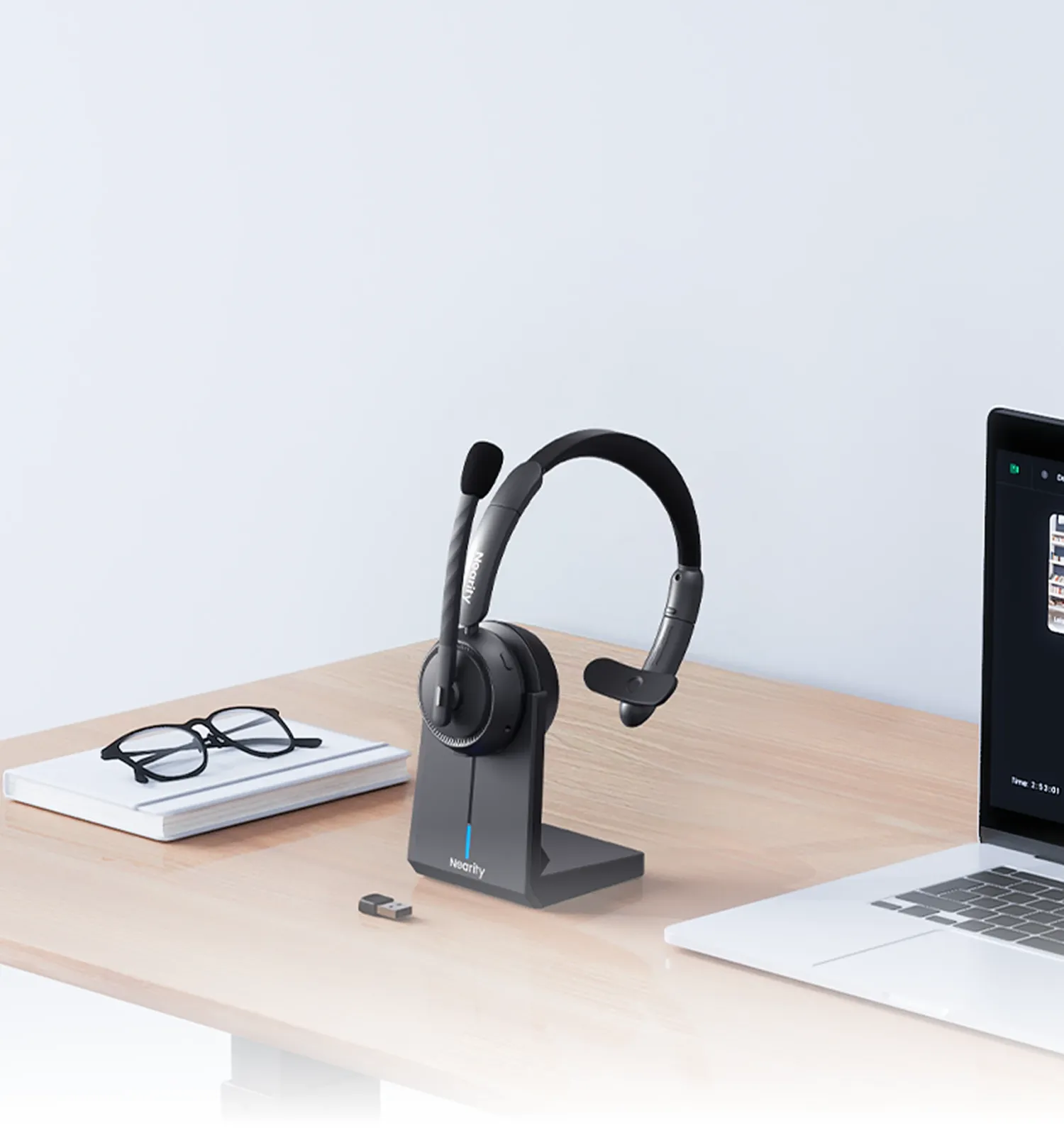 A professional single ear wireless headset with mic for pc placed on its charging dock next to a laptop running a video conference, showing a perfect home office setup with a one ear headphone with mic.