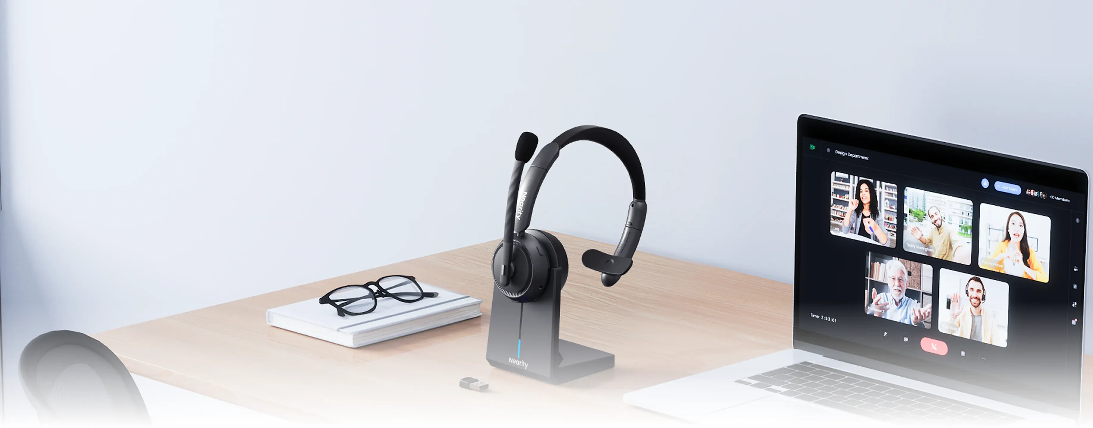A professional single ear wireless headset with mic for pc placed on its charging dock next to a laptop running a video conference, showing a perfect home office setup with a one ear headphone with mic.