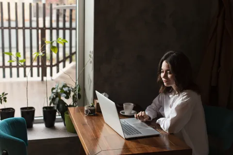 A woman sitting at a small wooden table in a brightly lit coffee shop, focused on her silver laptop. She has a white ceramic coffee mug next to her and is wearing a comfortable knit sweater, illustrating a modern remote work setting.