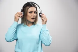 Blonde girl in a blue sweatshirt frowning while her headphones are not working